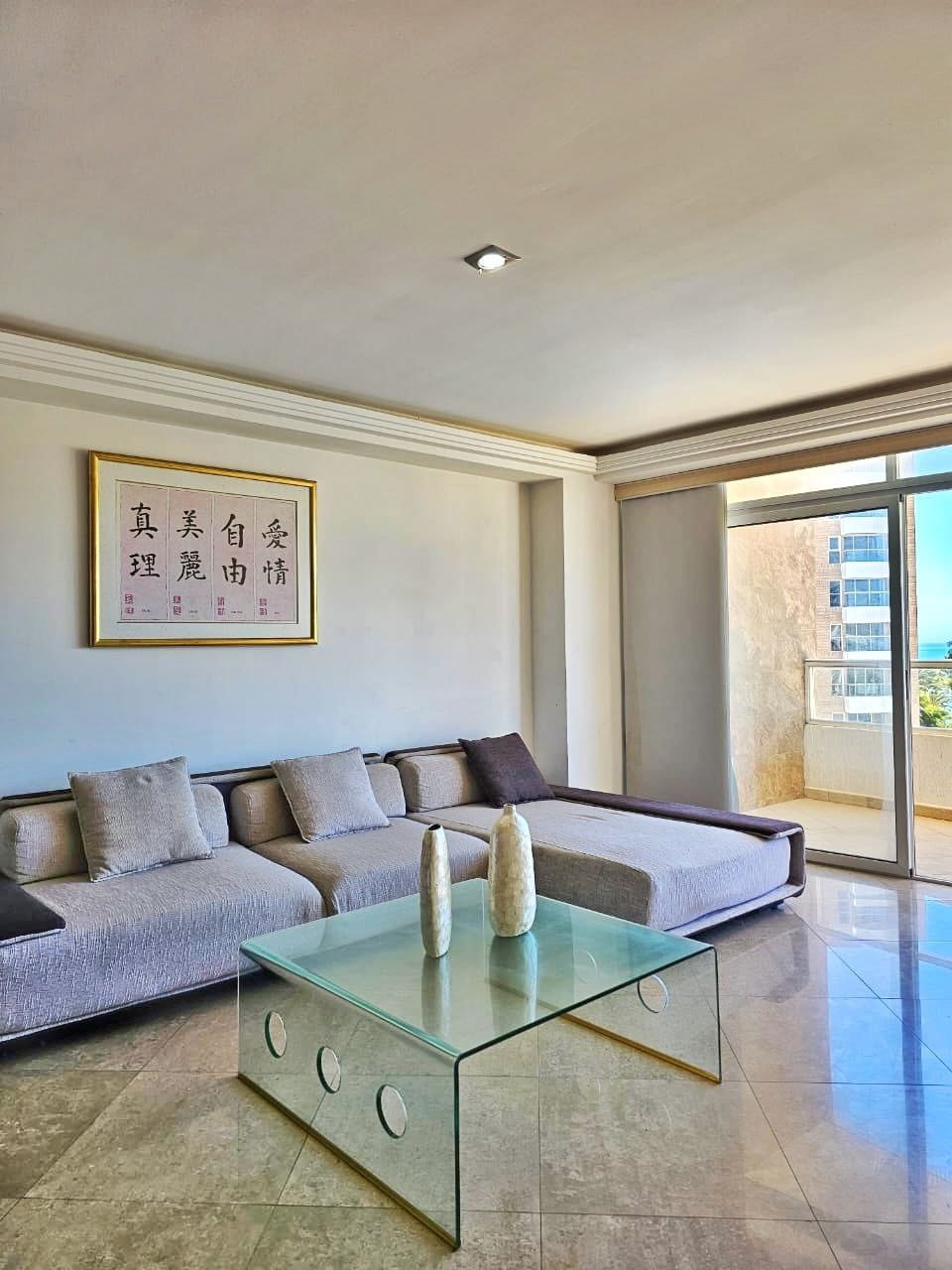 A spacious living area features a contemporary sofa adorned with neutral-colored cushions. A glass coffee table with decorative vases is positioned in front. Natural light filters in through large sliding doors, offering a view of the ocean beyond.