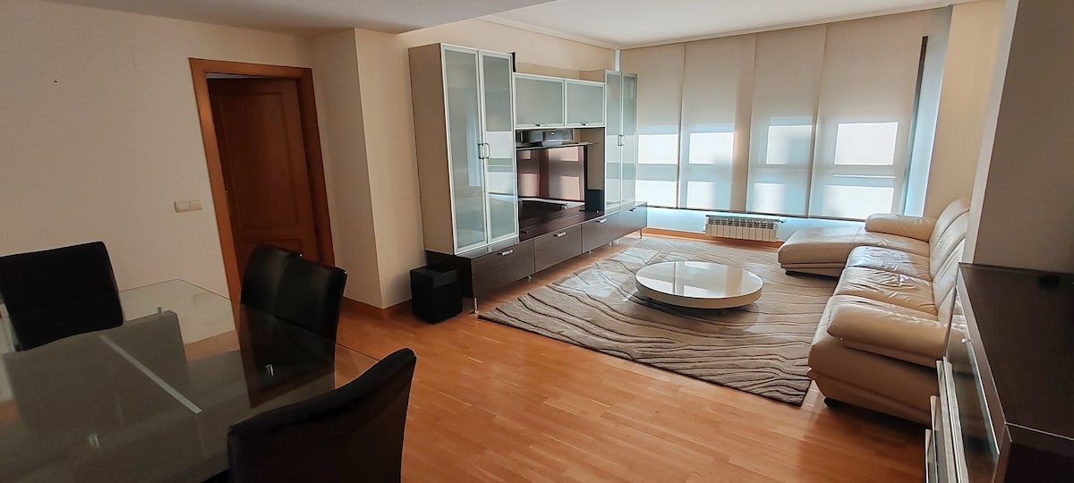 A bright living area features large windows allowing natural light to fill the space. A light-colored leather sofa and a circular coffee table are arranged on a textured area rug. A glass dining table with black chairs sits nearby, and a TV unit provides entertainment options.