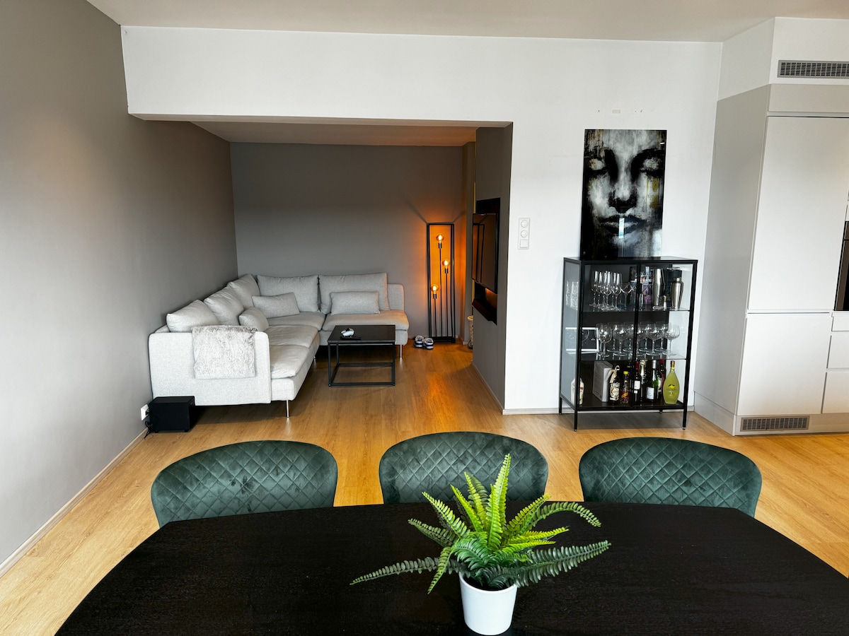 A spacious dining area is depicted, featuring a dark table surrounded by green chairs. In the background, a cozy seating arrangement is visible with a light-colored sofa. A modern cabinet displaying glassware stands against a wall, adding functional elegance to the space.