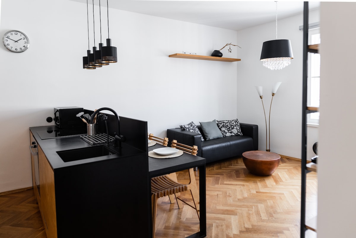 The open kitchen and living area feature modern furnishings, including a sleek kitchen island and a dark sofa with decorative pillows. A wood dining table is set for two, and light fixtures above add a contemporary touch. Natural light enhances the warm wooden flooring.