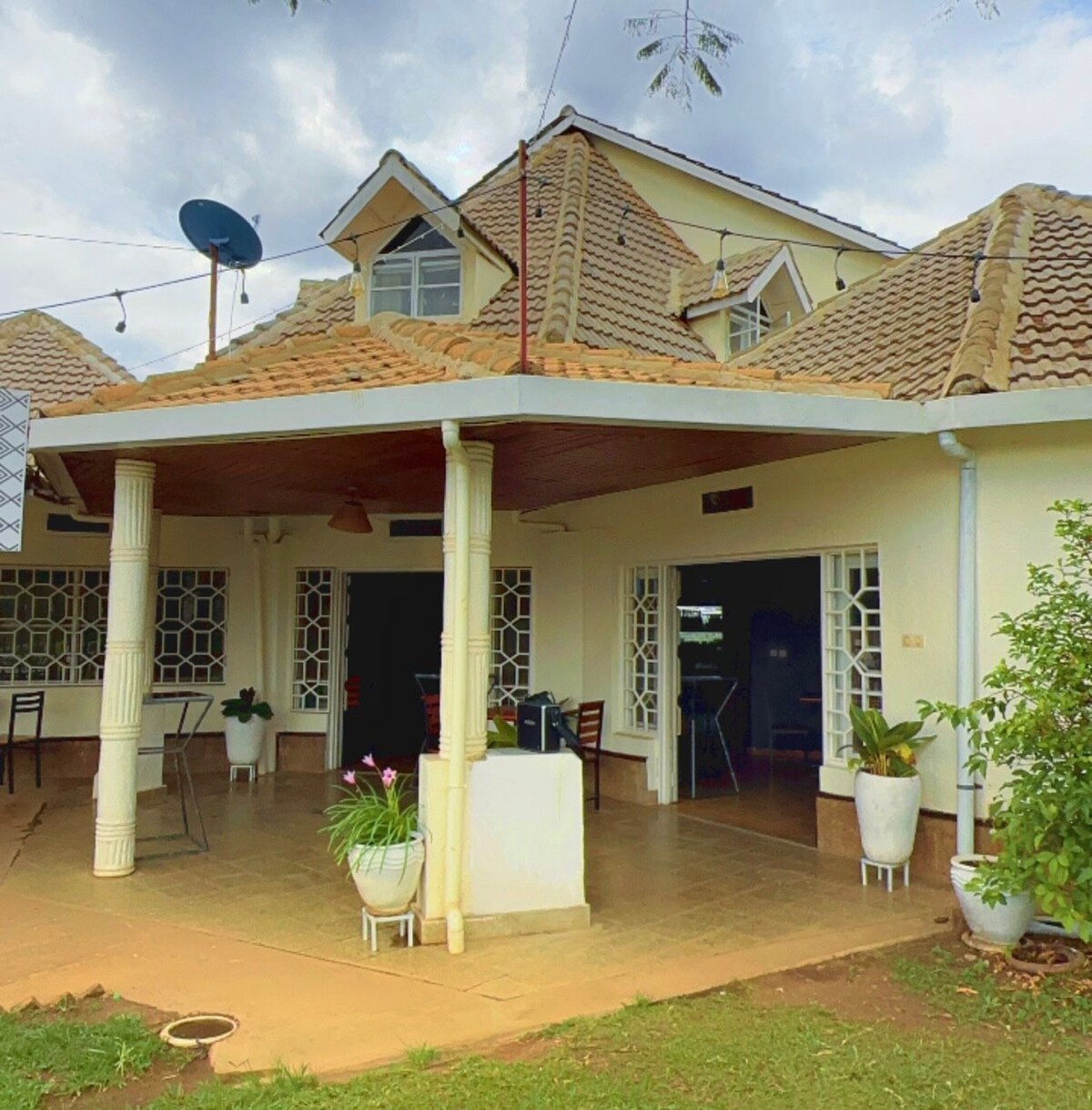 A charming exterior features a sloped roof and inviting porch, framed by white columns. Glass panel doors lead to an open area with seating arrangements. Potted plants add a touch of greenery, while the surrounding landscape provides a serene setting.