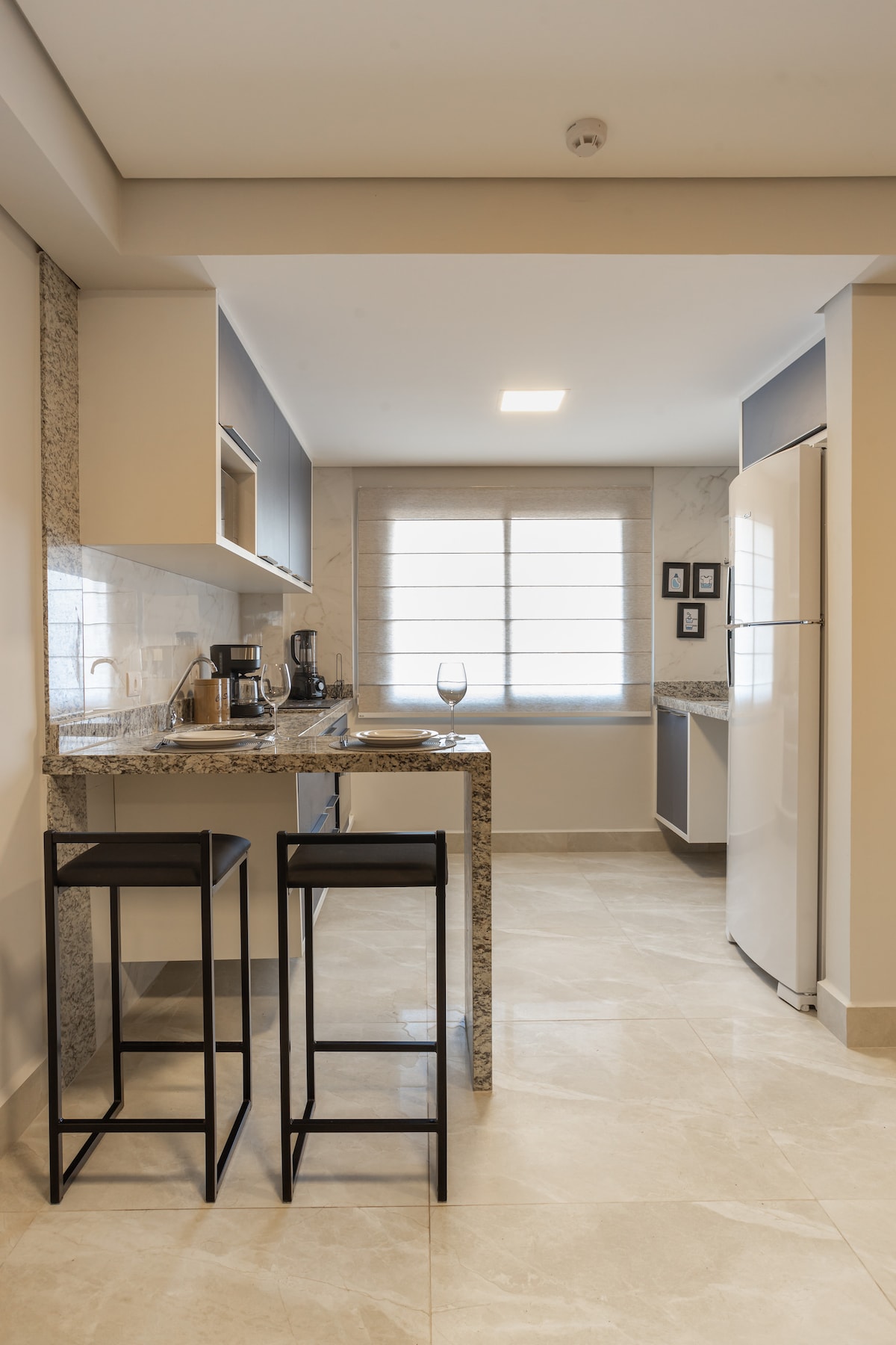 A well-equipped kitchen area is visible, featuring modern appliances and a granite countertop. Two high-backed stools are positioned at a small bar, offering a space for casual dining. A large window allows natural light to fill the room, while light-colored cabinets provide a clean look.