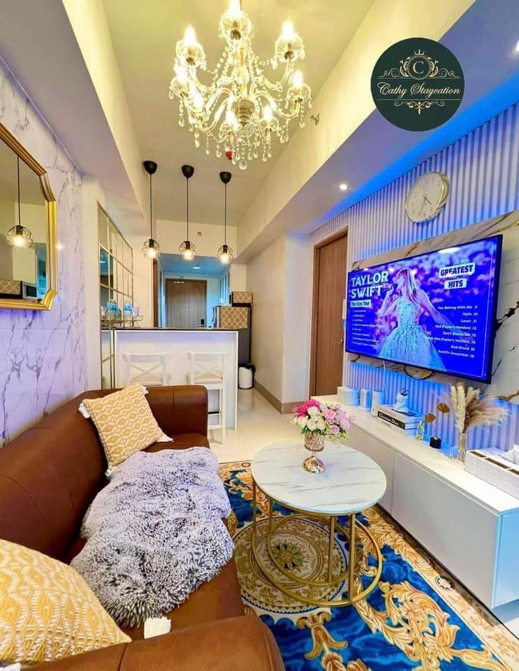 A stylish living area features a brown sofa adorned with textured cushions, complemented by a round marble coffee table. A statement chandelier hangs above, while a wall-mounted TV displays entertainment options. Soft lighting and a decorative rug enhance the cozy ambiance.