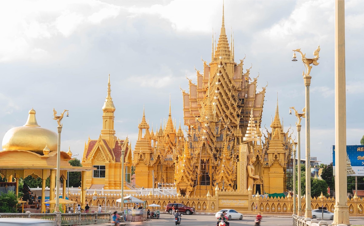 An ornate temple complex features golden spires and intricate architectural details, showcasing traditional Thai design. Visitors can be seen on motorcycles in the foreground, with the temple serving as a striking backdrop against a cloudy sky.