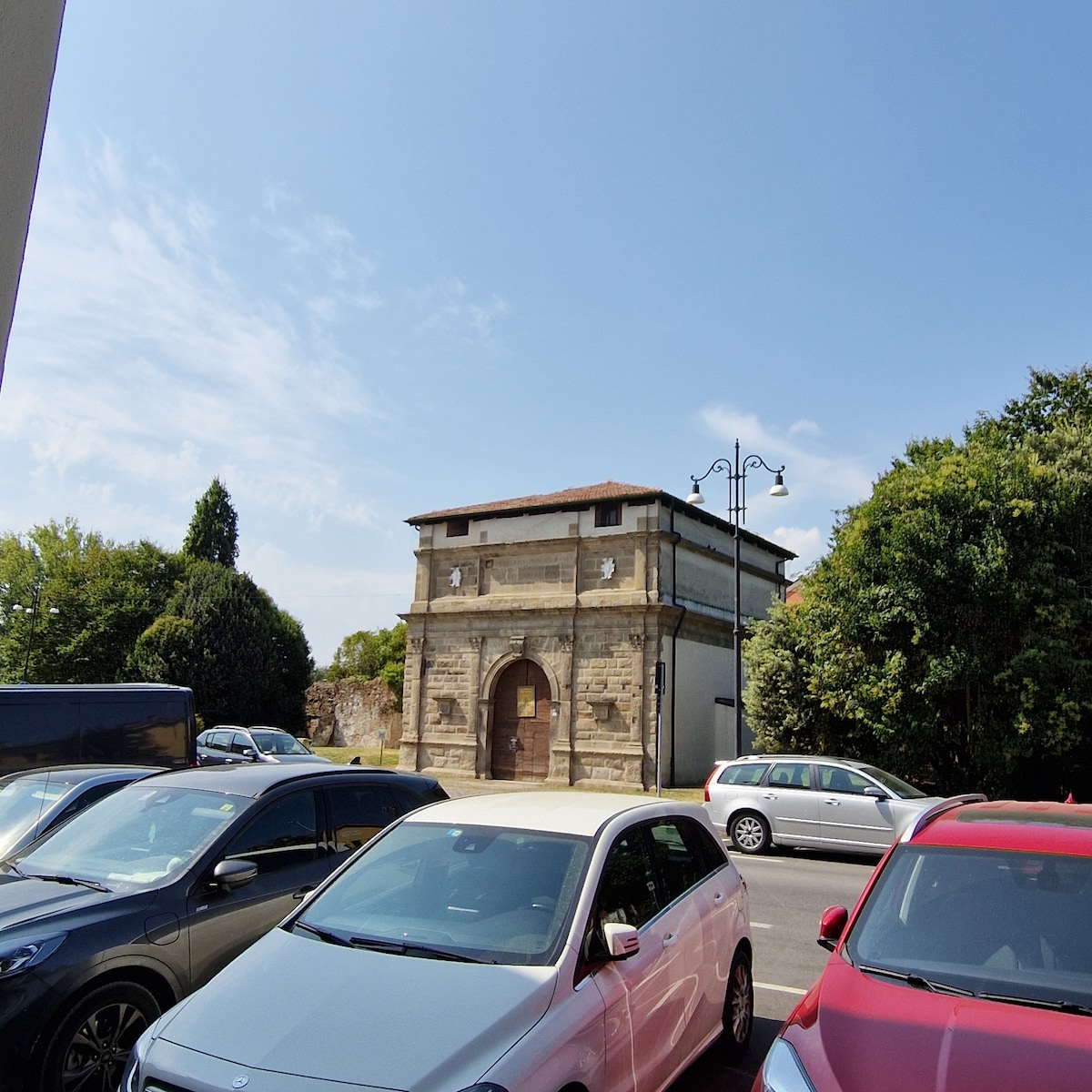 The image shows a historical building, characterized by its stone façade and arched entrance. Surrounding vehicles are parked closely, along with greenery in the background. A clear blue sky and scattered clouds create a bright atmosphere.