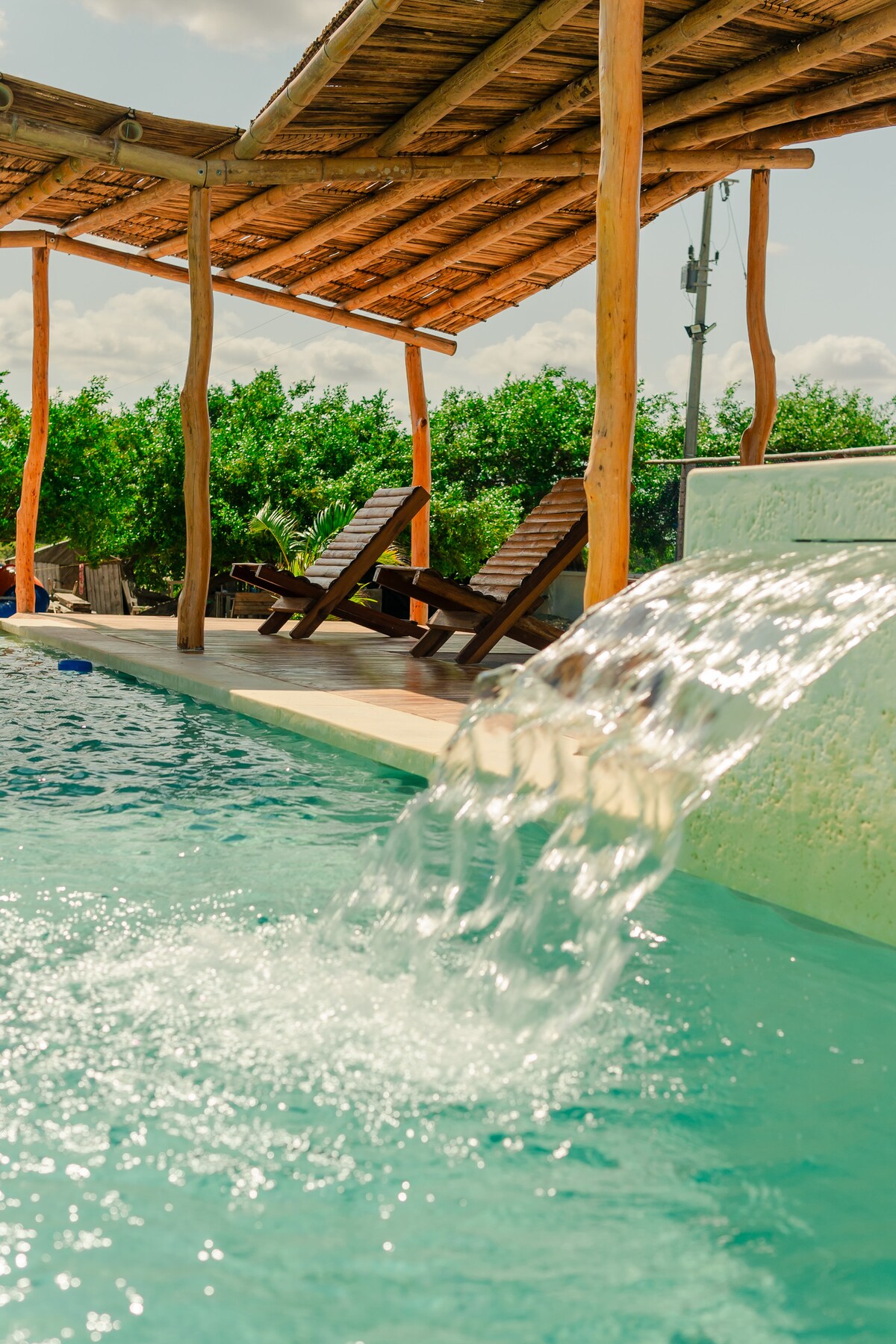 A swimming pool is highlighted by a waterfall feature, with water cascading gently into the turquoise waters. Wooden lounge chairs are positioned nearby, offering a place to relax under a thatched roof gazebo. Lush green trees frame the scene, contributing to a serene atmosphere.