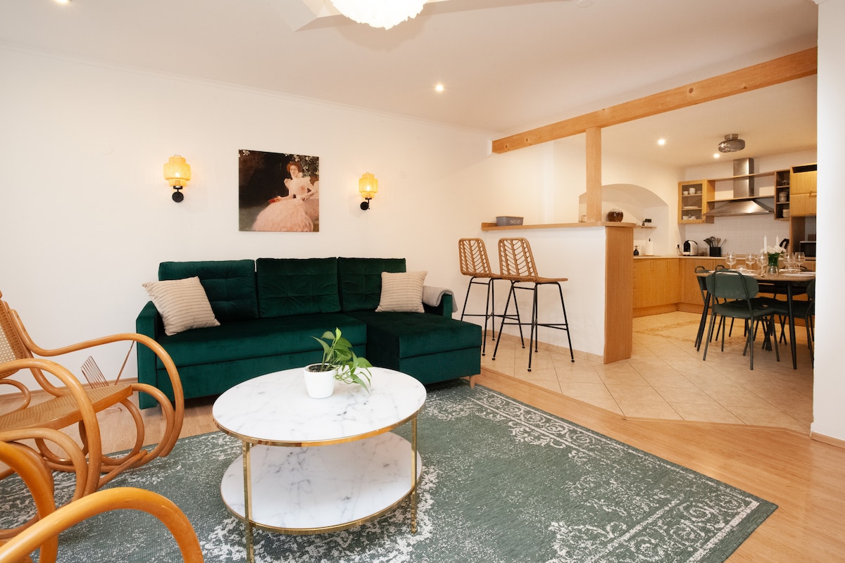 A spacious living area features a large green sectional sofa and a round marble coffee table. Soft lighting is provided by wall sconces. In the background, the open-concept kitchen and dining area are visible, showcasing wooden cabinetry and modern appliances.
