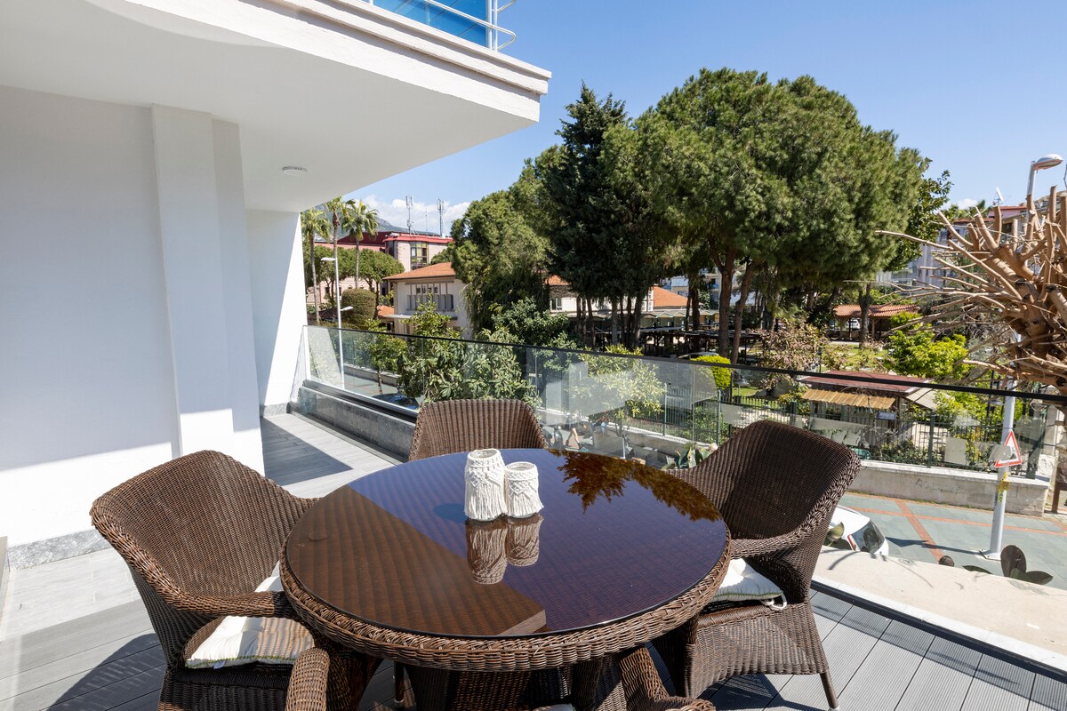 A terrace featuring a round glass-top table surrounded by four wicker chairs is visible. The area is adorned with greenery and trees in the background, providing a serene outdoor setting. Natural light enhances the inviting atmosphere.