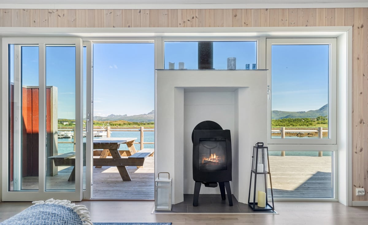 A cozy living area is highlighted by a modern fireplace positioned between large glass doors that open to a scenic patio. Natural light enters through the unobstructed views of the waterfront and the surrounding landscape, enhancing the peaceful ambiance.
