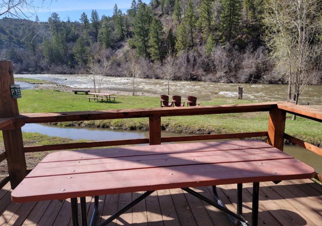 A sturdy picnic table is set on a wooden deck, overlooking a flowing river. Green grass surrounds the area, with several red chairs positioned nearby. Dense trees line the riverbank, creating a natural backdrop that enhances the outdoor dining experience.