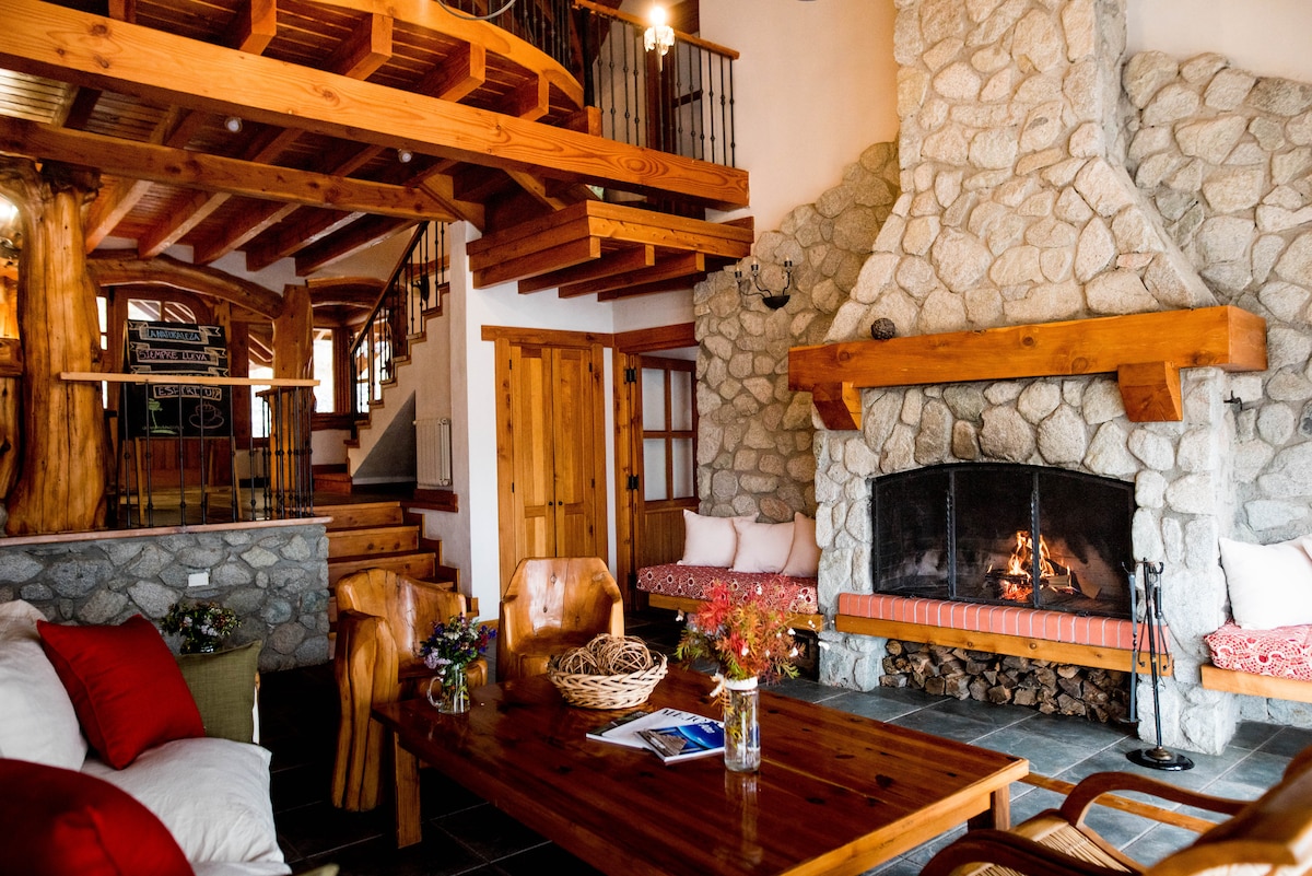 A rustic living area is framed by wooden beams and stone walls. A large fireplace serves as a focal point, surrounded by comfortable seating. Natural light filters through windows, highlighting the wooden furniture and a cozy atmosphere that invites relaxation.
