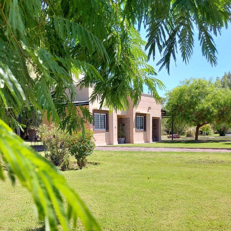 The exterior of a charming pink building is framed by lush green leaves, revealing a welcoming front porch. Surrounding greenery and well-maintained lawns enhance the serene environment. Windows provide a glimpse of the interior, contributing to the overall inviting appearance.