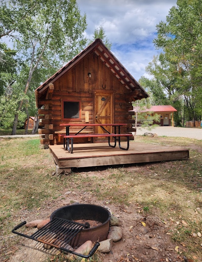 A rustic cabin constructed from logs features a wooden deck with a picnic table. A fire pit is placed nearby, surrounded by stones. Green trees and partly cloudy skies enhance the natural setting, providing a peaceful outdoor atmosphere.