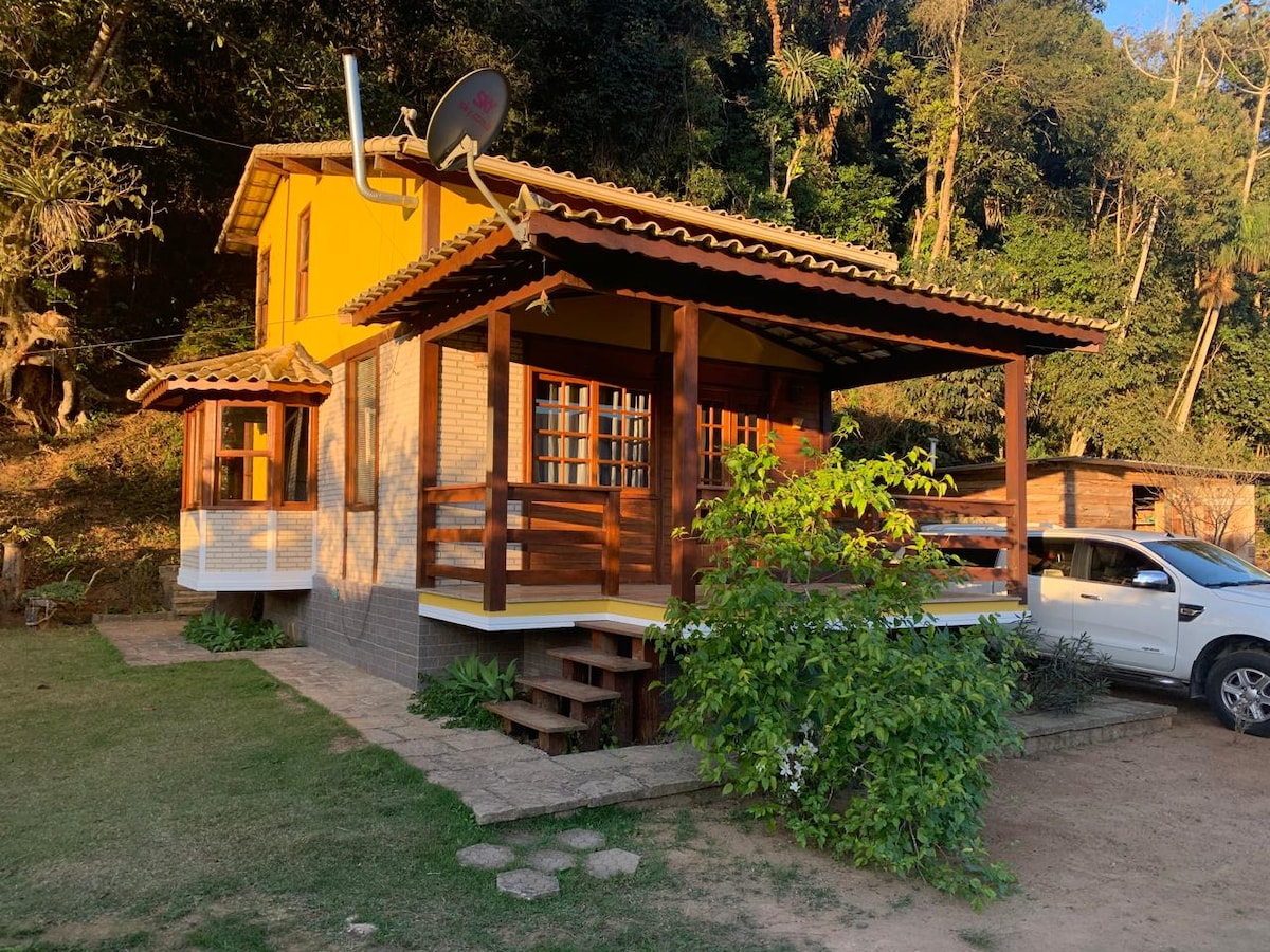 A charming two-story chalet is surrounded by lush greenery, featuring a wrap-around porch with wooden railings. A set of stairs leads to the entrance, while large windows allow natural light to illuminate the interior. A parked vehicle is visible beside the building.