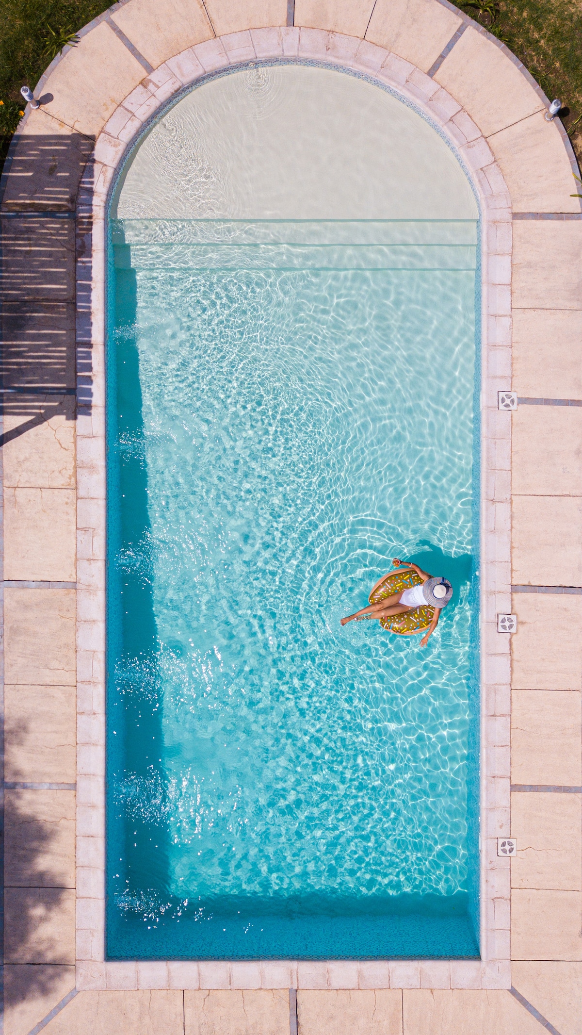 An aerial view of a rectangular pool is displayed, surrounded by pale stone decking. Light reflects off the clear water, creating a serene atmosphere. A person is seen floating on the surface, adding a touch of life to the tranquil setting.