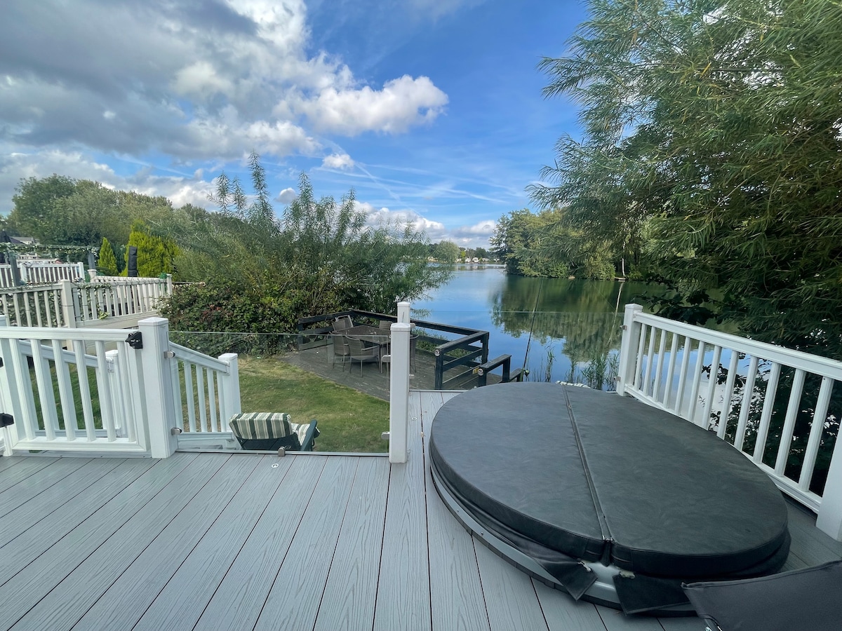 A scenic view from a private deck overlooks a calm lake, framed by lush greenery. A circular hot tub is visible, with a safety cover resting on top. Comfortable seating is positioned nearby, offering a perfect spot to enjoy nature.