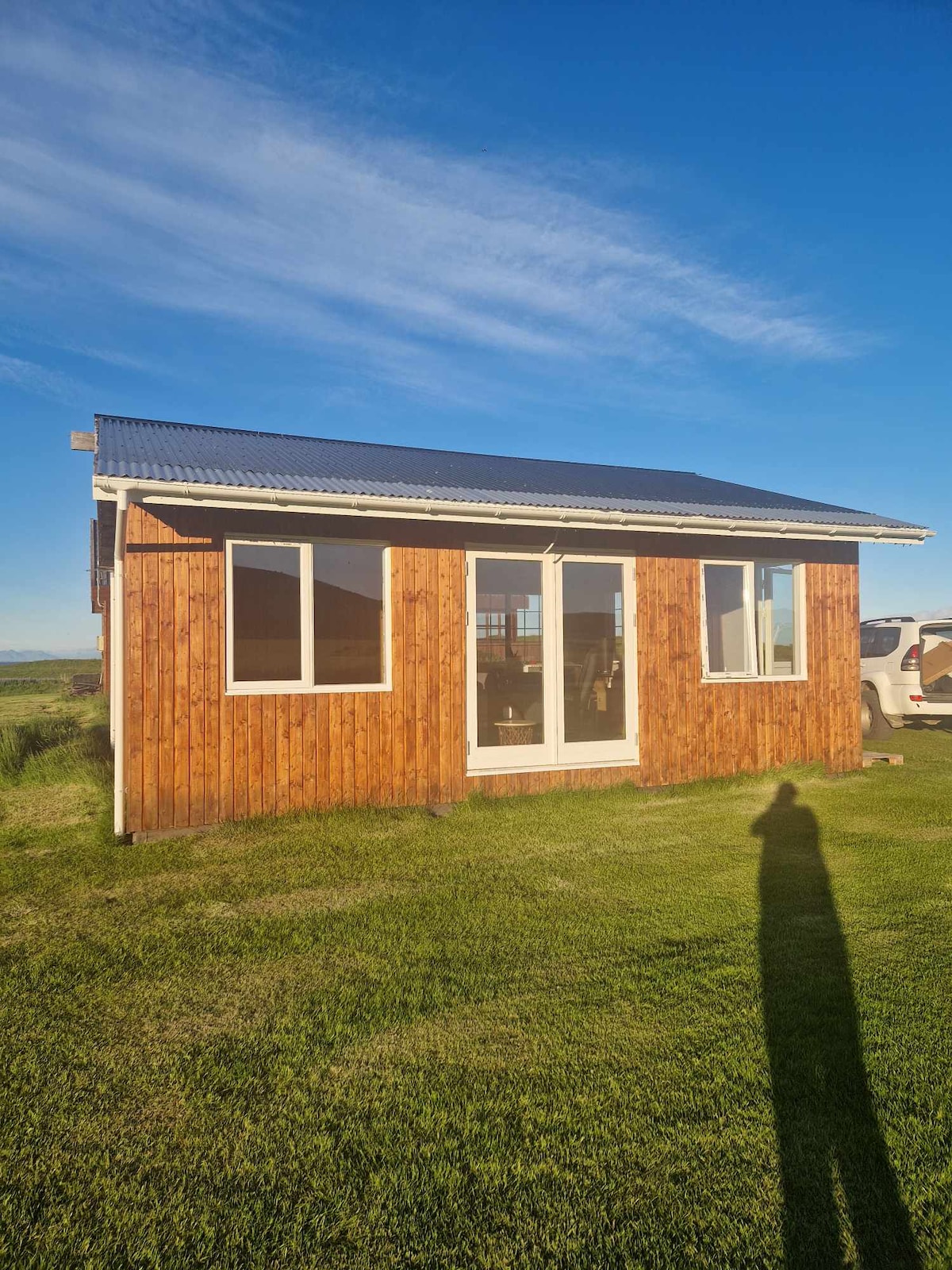 A wooden cottage exterior is shown under a clear blue sky. Large windows allow for natural light, while a white door leads into the inviting space. A well-maintained grassy area surrounds the building.