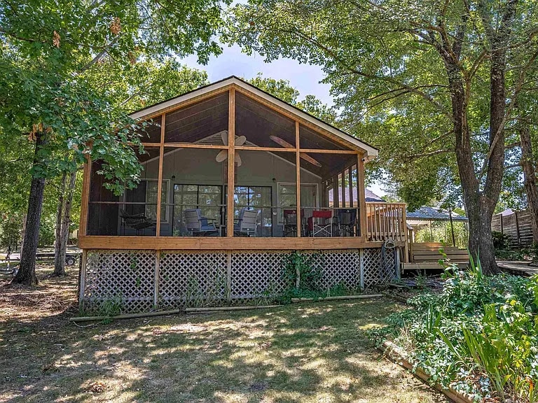 A screened back patio is shown, featuring a wooden structure with a sloped roof and multiple seating arrangements. Surrounding trees provide shade, while a deck leads to the entrance. The patio offers views of the outdoor space, creating a serene environment for relaxation.