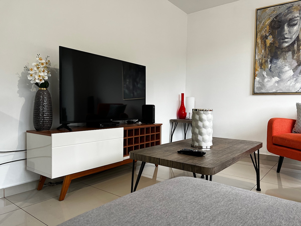 A modern living area is displayed, featuring a large flat-screen television, a stylish wooden coffee table, and a sleek white media console. A vibrant orange chair and decorative elements, including a vase and abstract art, add visual interest to the neutral-colored space.