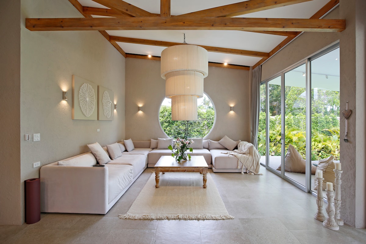 A spacious living room features a large, light-colored sectional couch and a round window that offers views of the surrounding jungle. A coffee table and decorative elements adorn the area, while natural light fills the space through expansive glass doors.