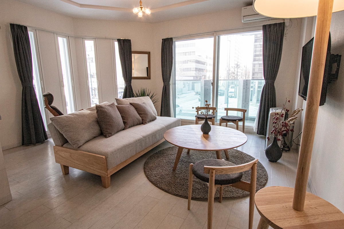 A spacious living area features a comfortable sofa and a round wooden coffee table atop a textured rug. Large windows allow natural light to fill the room, with views of the outside. Simple wooden chairs and decorative plants add a touch of style.