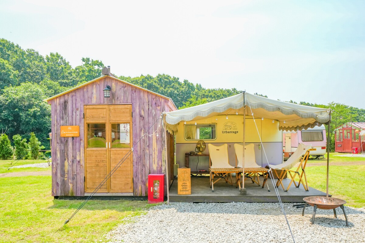 A charming wooden cabin is positioned alongside a covered area featuring lounge chairs. A gravel pathway leads to the entrance, with lush green grass surrounding the structures. The backdrop is filled with dense trees, contributing to a tranquil outdoor setting.