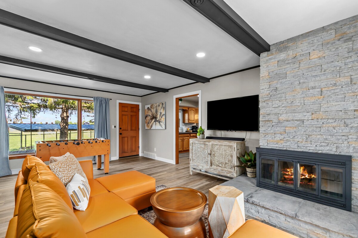 A bright living area features a spacious orange sectional sofa facing a stone fireplace. A large window offers views of the serene outdoor space. A foosball table and wooden door lead to the kitchen, enhancing the inviting atmosphere.