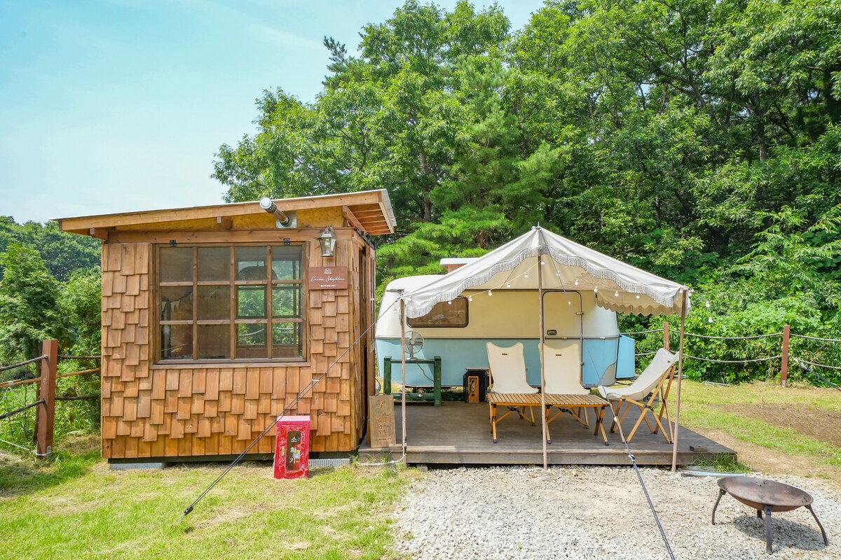 A charming cabin with a wooden exterior is accompanied by a vintage caravan under a white canopy, situated on a wooden deck. The surrounding area features lush greenery, providing a serene backdrop. Two comfortable chairs are arranged on the deck, inviting relaxation.
