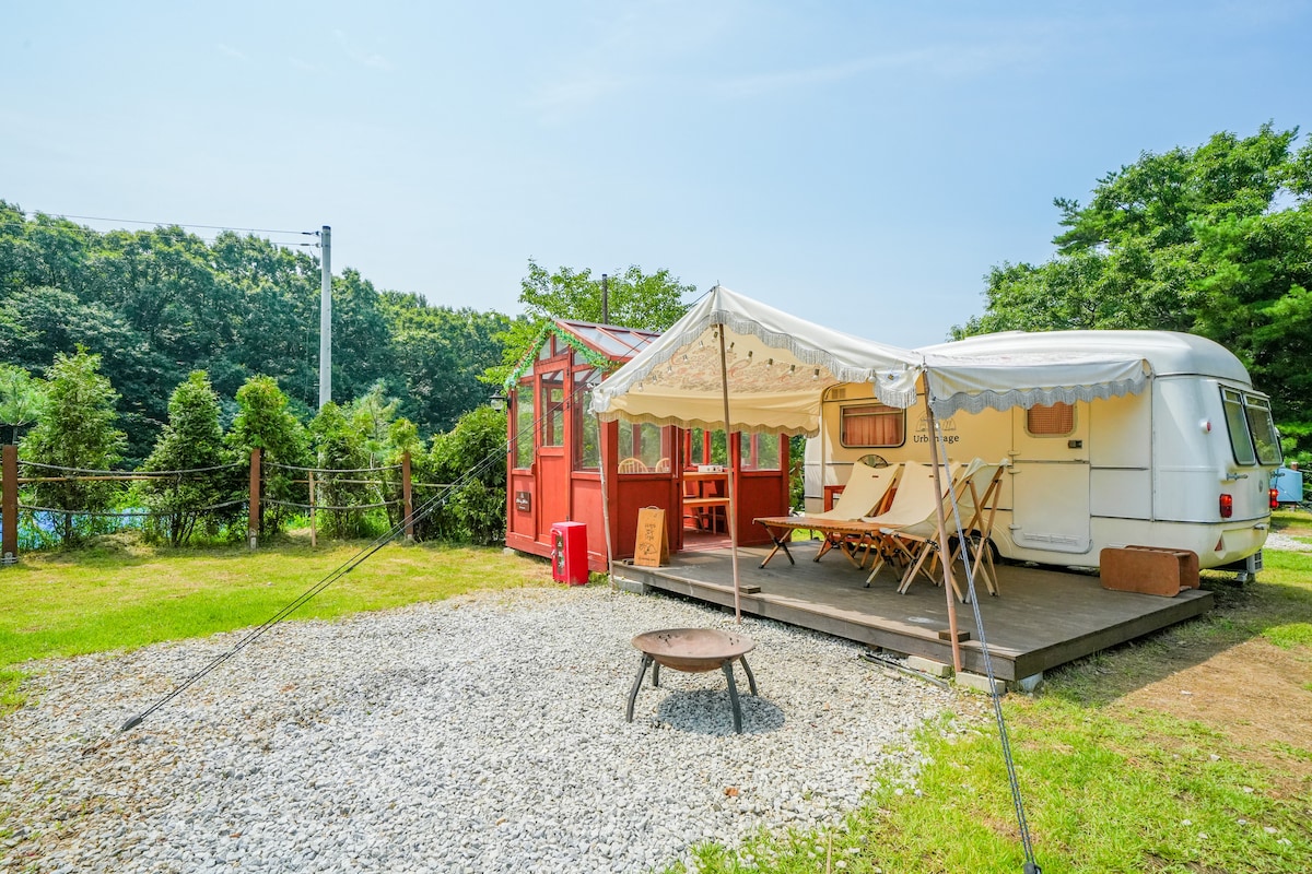 A vintage caravan is positioned on a gravel patio, surrounded by lush greenery. A red cabin with large windows sits nearby, while two lounge chairs are placed on the deck under a white canopy. A fire pit is set on the grass, providing a cozy outdoor gathering space.