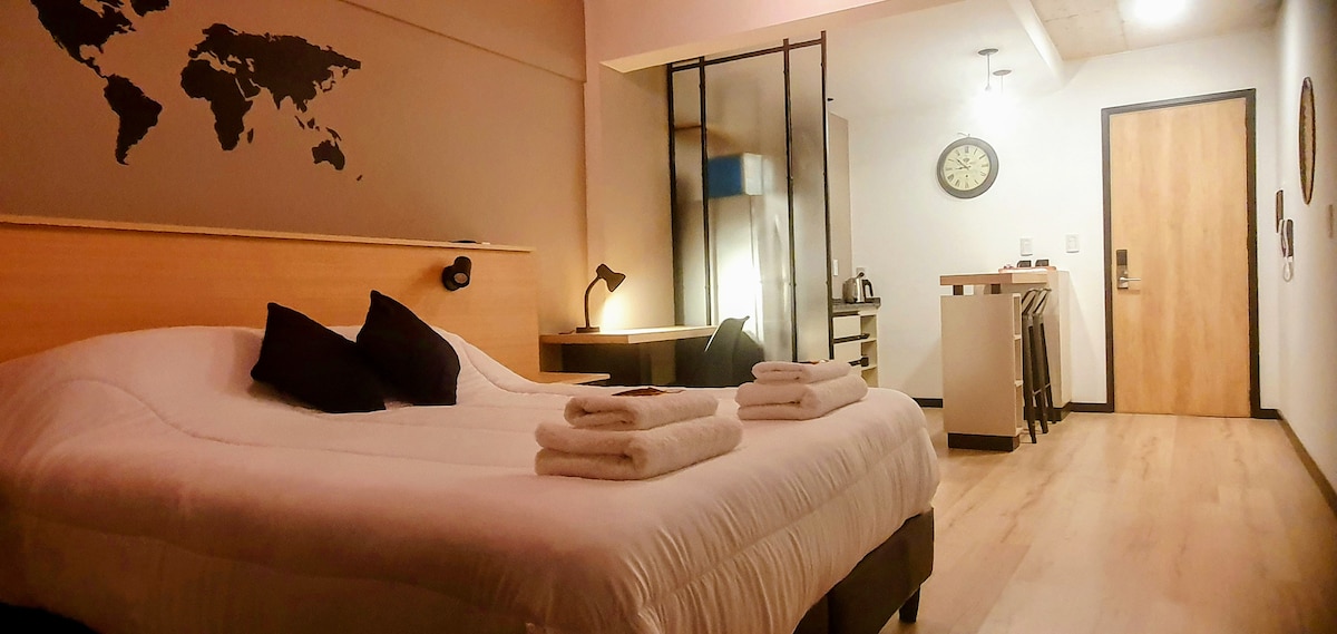 A modern studio apartment is presented with a spacious bed featuring crisp white linens and decorative pillows. A wooden desk and chair are positioned near a glass partition, while a kitchenette area, complete with bar stools, is visible in the background. A world map decorates the wall.