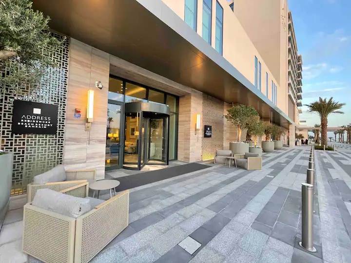 The entrance of the Address Beach Resort Residence is framed by modern architecture. Seating areas with contemporary furniture are positioned beside a glass revolving door. Palm trees and a light stone pathway enhance the welcoming exterior, while the building's facade is lined with large windows.