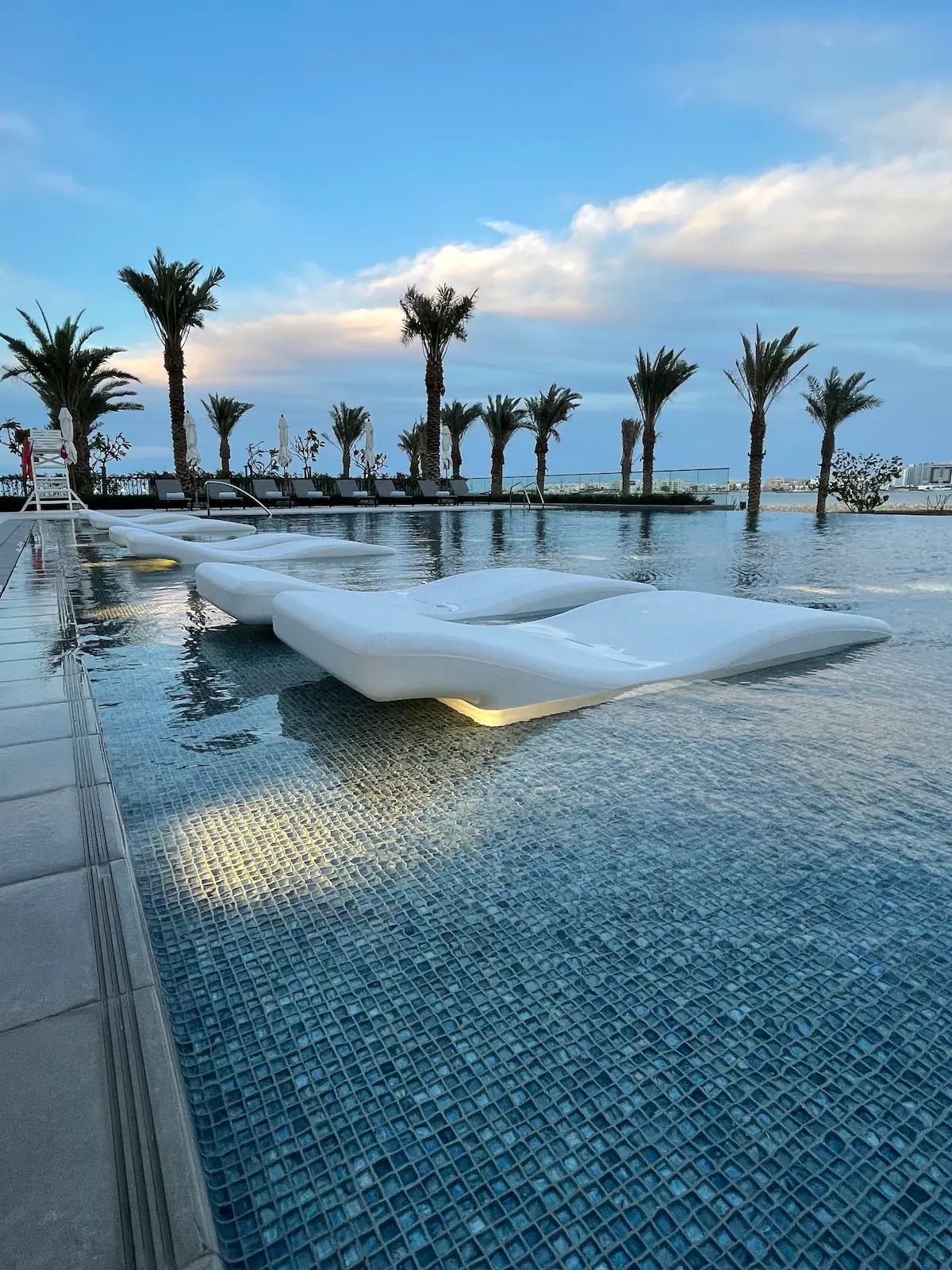 A serene pool area is shown, featuring a stylish white lounge platform partially submerged in the water. Palm trees line the pool's edge, and lounge chairs are positioned for relaxation, all under a sky with soft, drifting clouds.