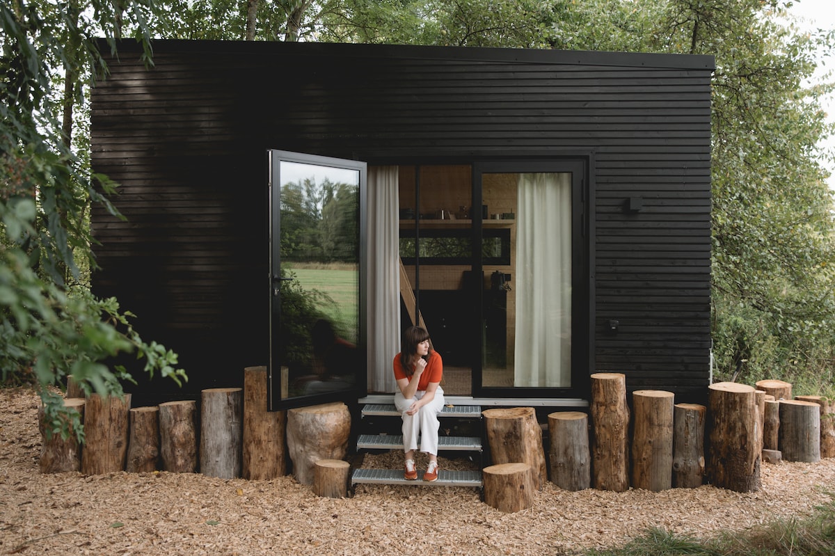 The entrance of the tiny house is marked by a black wooden facade, complemented by large glass doors that open to a natural setting. Log seating surrounds the area, offering a rustic touch. A green field is visible in the background, enhancing the tranquil atmosphere.