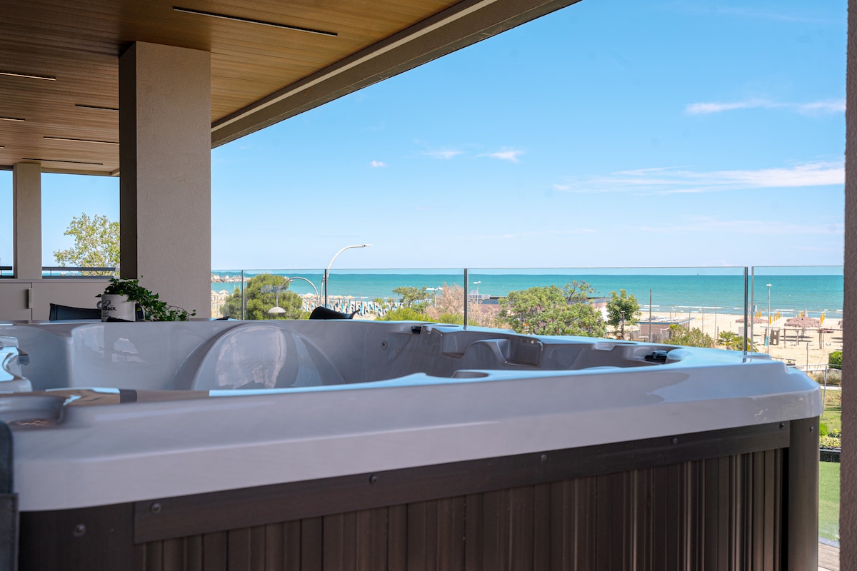 A hot tub is positioned on a balcony with a clear view of the ocean. The surrounding area features greenery and a pathway leading to the beach. The horizon showcases the serene blue waters under a bright sky.