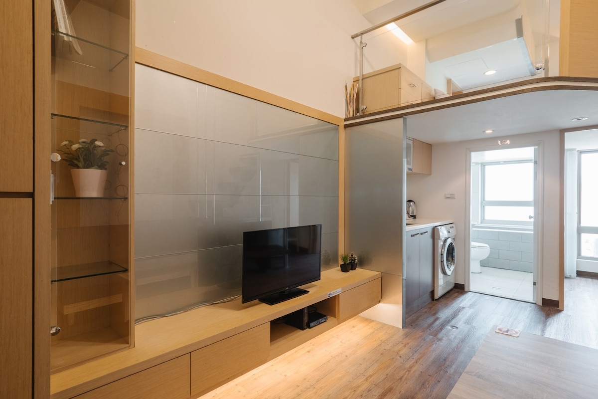 An open living space is highlighted by a flat-screen television mounted on a wooden entertainment unit. Natural light fills the room through large windows, enhancing the modern design features. A washing machine is visible in the background, providing convenient laundry access.