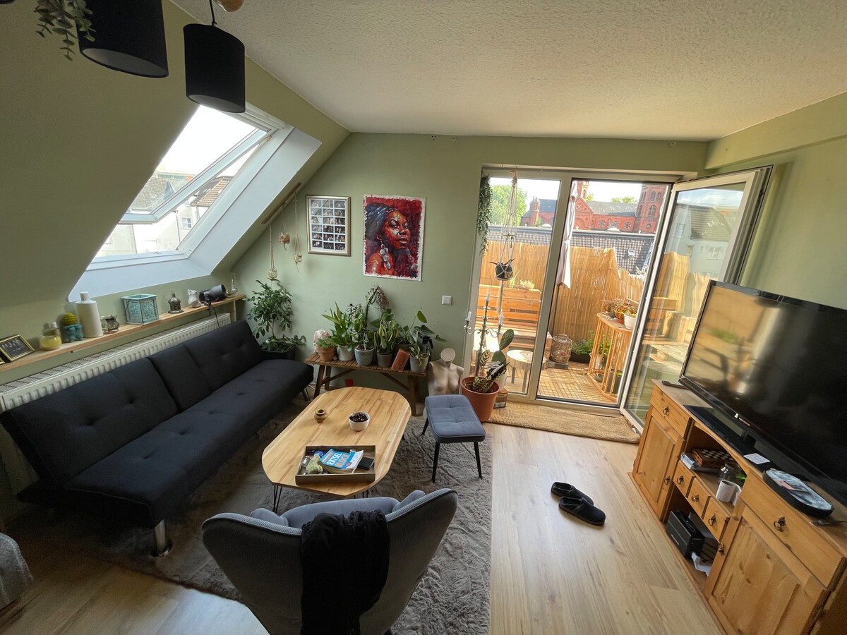 A bright living space features a spacious black sofa and a wooden coffee table surrounded by various potted plants. A comfortable chair sits nearby. Natural light pours in through large windows and a sliding glass door, leading to a small outdoor balcony area.