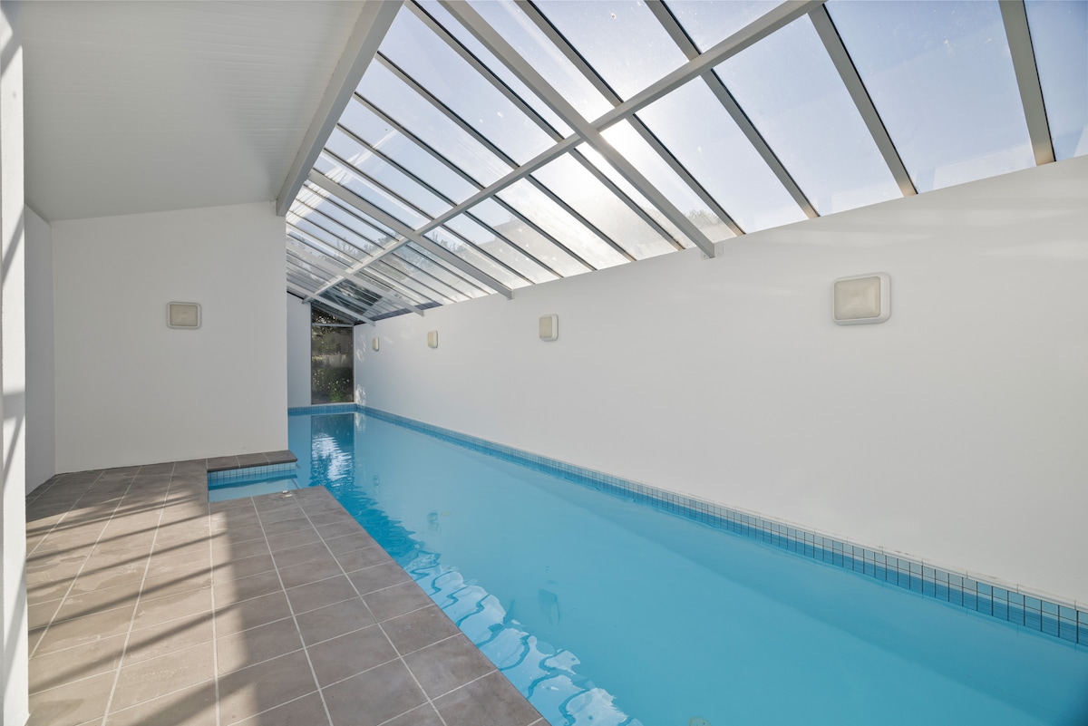 An indoor pool area is displayed, featuring a long, narrow pool with clear blue water. Sunlight enters through a transparent roof, highlighting the smooth, tiled edges. Minimalistic white walls frame the space, with soft lighting fixtures positioned along the sides.