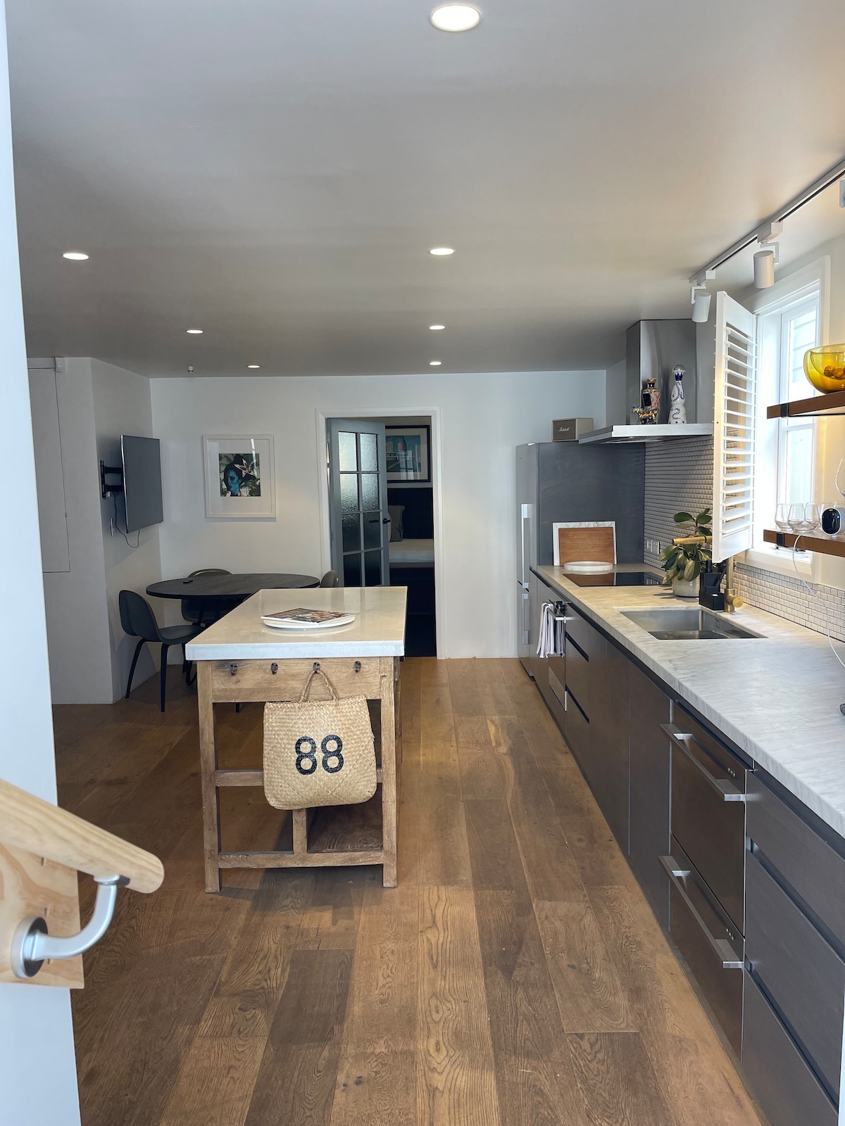 A modern kitchen features sleek cabinetry and a spacious island with a wooden base and a countertop. Recessed lighting illuminates the area, highlighting the open layout. An adjacent dining space with a small table and chairs is visible, creating a cohesive atmosphere.
