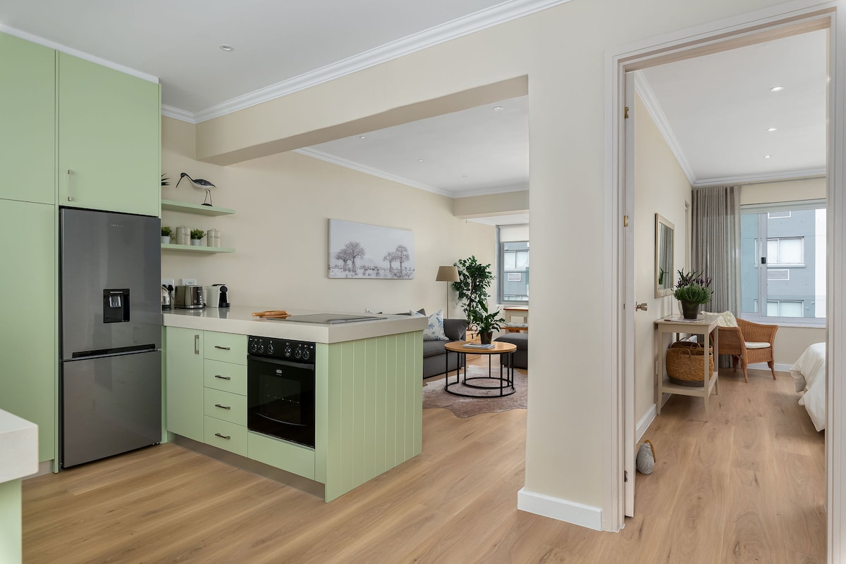 An open-plan kitchen and lounge area is presented, featuring mint-green cabinetry and modern appliances. A spacious island countertop complements a subtle dining area. Natural light illuminates the inviting lounge, furnished with a comfortable sofa, while a bedroom is visible through the doorway, providing a cozy atmosphere.