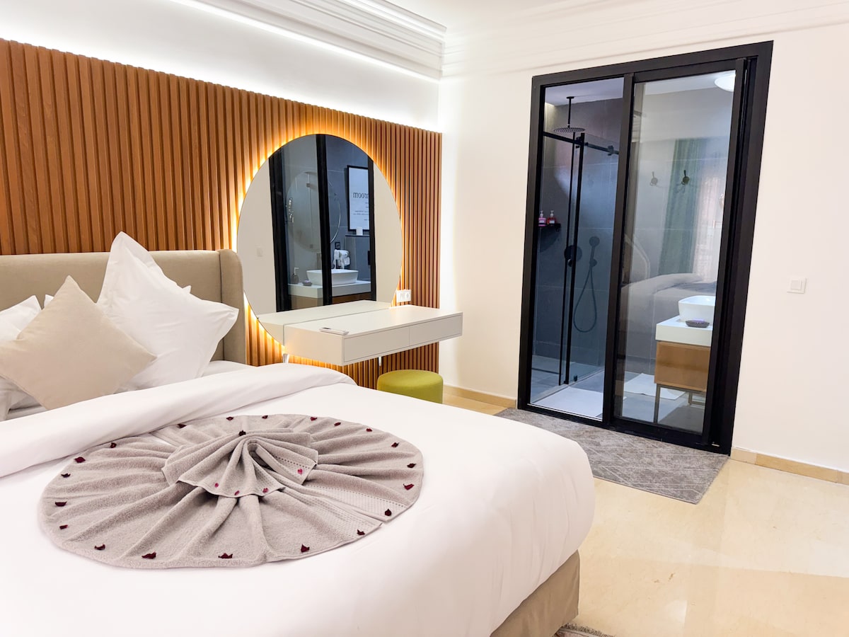 A well-appointed bedroom features a spacious bed with an elegant blanket draped artistically over it. A round mirror with soft lighting complements the wooden paneling. A glass door leads to a modern bathroom, visible in the background, showcasing sleek design elements.