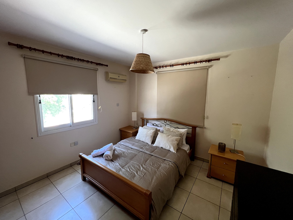 A cozy bedroom features a wooden bed with neutral bedding and multiple pillows. Two bedside tables are positioned beside the bed, each topped with a lamp. Natural light enters through a window adorned with beige blinds, complemented by a stylish hanging light fixture.