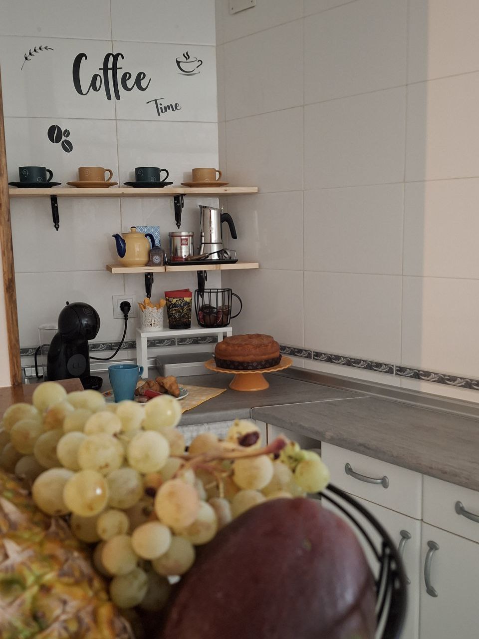 A neatly organized kitchen corner features shelves with various cups and coffee-making equipment. A fruit basket filled with grapes, pineapple, and other fruits is prominently displayed in the foreground. A cake on a stand sits on the countertop, contributing to a welcoming ambience.