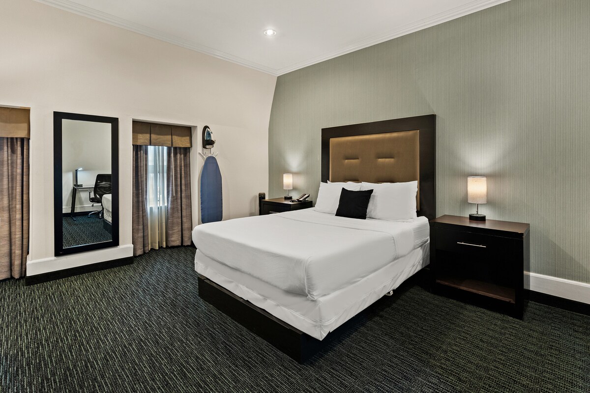 A spacious sleeping area features a large bed with a light-colored duvet and neutral-toned walls. Nightstands with lamps are positioned on either side of the bed. A full-length mirror and an ironing board are visible, along with a workspace illuminated by natural light from the window.