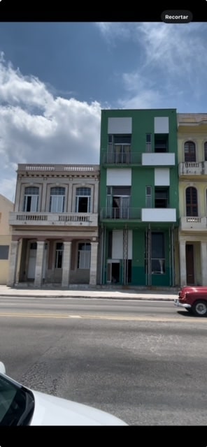A street view showcases a two-story building in a vibrant green color, flanked by traditional architecture. The modern structure features large windows and a balcony, while the neighboring buildings exhibit classic decorative elements. The sky is partly cloudy, adding a dynamic backdrop to the scene.
