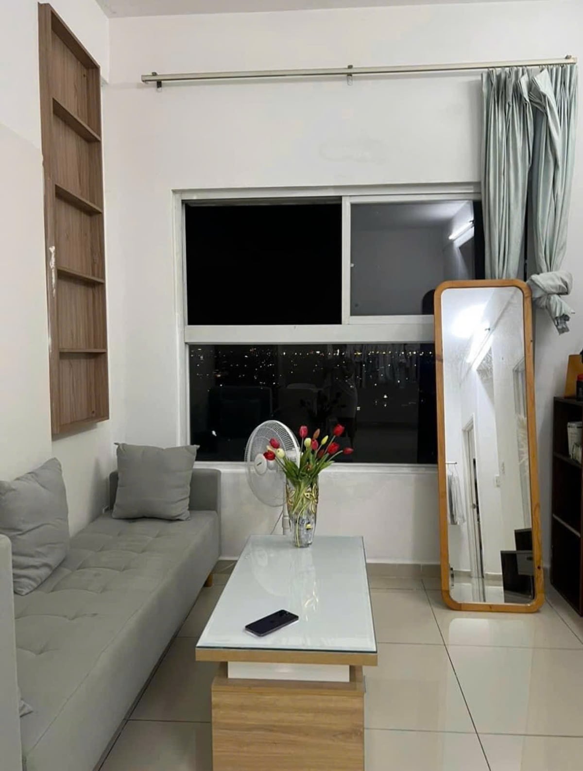 A living space features a gray sofa arranged against a light-colored wall, complemented by a wooden shelf. A large mirror leans against one wall, and a glass coffee table displays a vase with fresh flowers. A fan is positioned nearby, and a window offers a view of city lights.