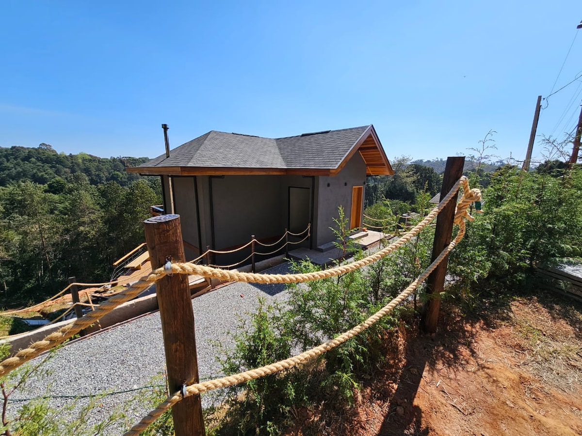 A charming house is pictured, nestled in a scenic landscape. Gravel pathways lead to the entrance, framed by rustic wooden railings entwined with rope. Lush greenery surrounds the property, complementing the clear blue sky and mountain views in the background.