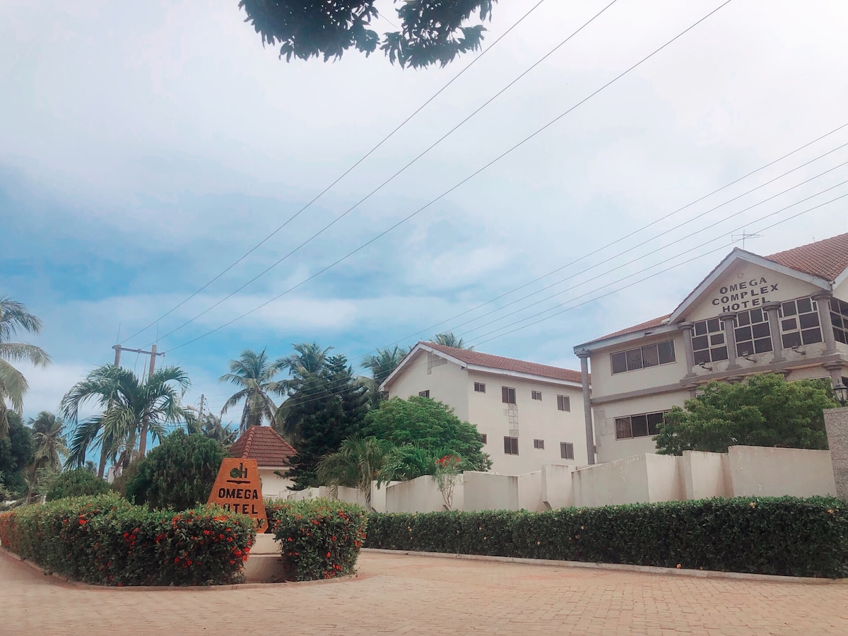 The exterior of Omega Complex Hotel is displayed, featuring a mix of architecture with a distinct sign prominently placed at the entrance. Lush green plants and low hedges line the paved pathway leading to the hotel, framed by palm trees under a cloudy sky.