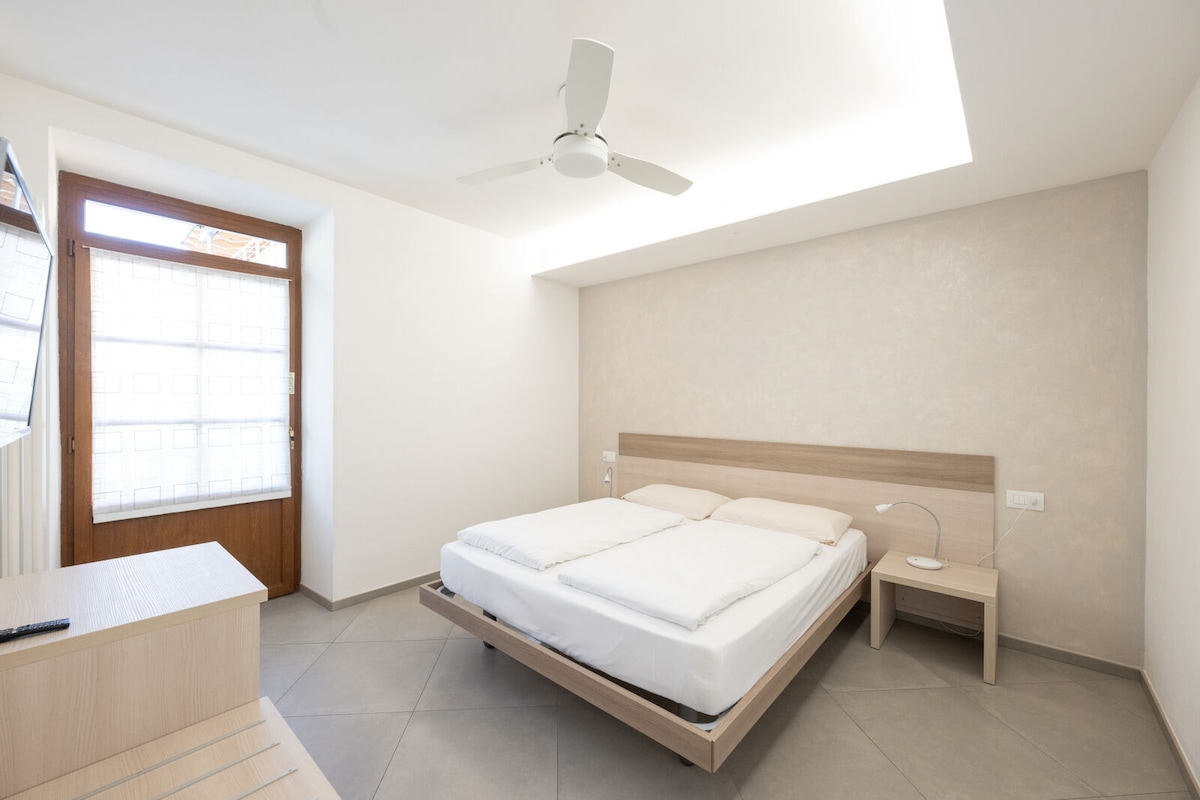 A bright and airy bedroom is seen, featuring a spacious bed with light bedding. A ceiling fan is installed above, while a large window allows natural light to fill the room. A minimalist nightstand and dresser complement the neutral color palette.