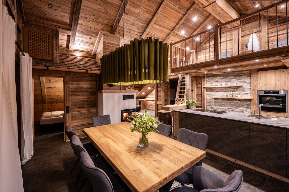 A spacious dining area features a large wooden table surrounded by upholstered chairs. The room is adorned with natural wood elements and modern lighting. A well-equipped kitchen with stone accents is visible, along with a cozy fireplace, enhancing the inviting atmosphere.