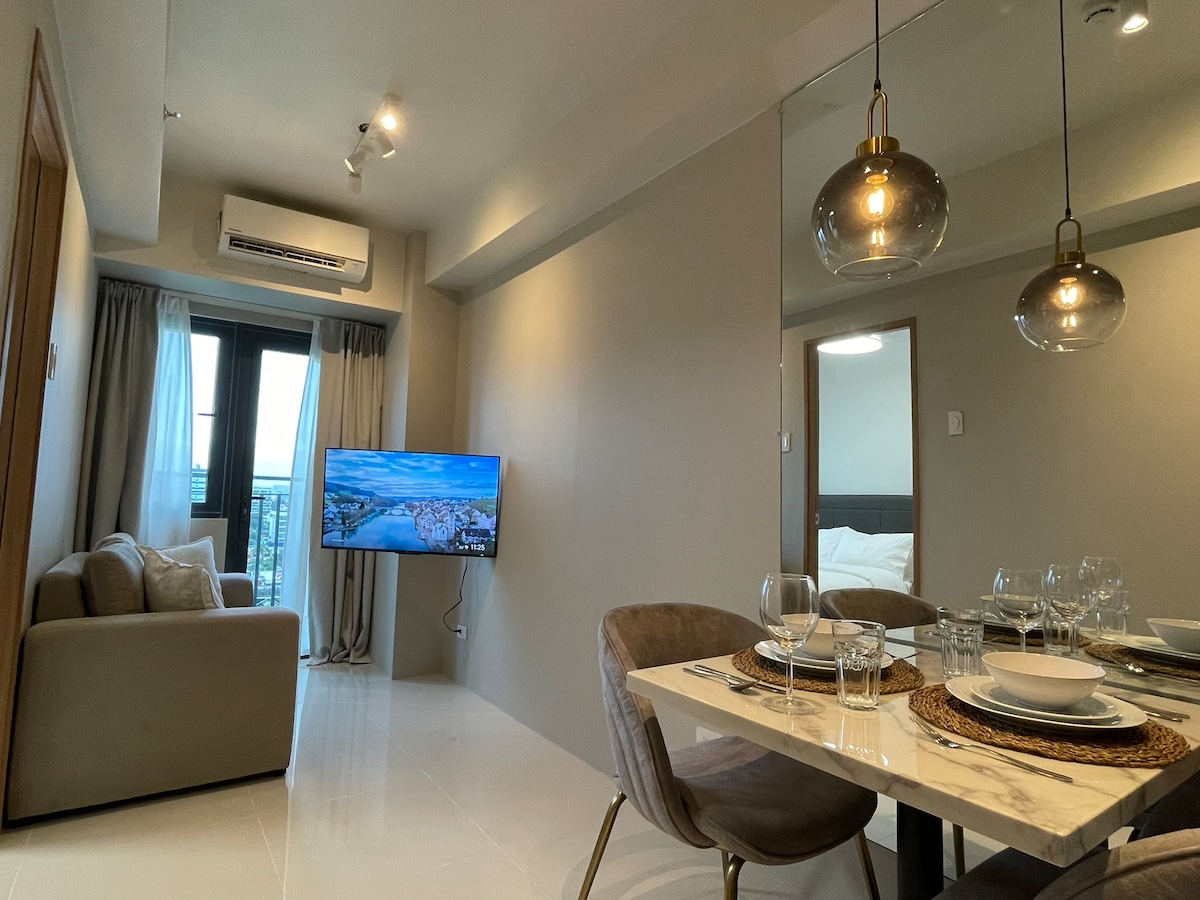 The living area features a cozy sofa and a 43-inch flat-screen TV, providing an inviting space for relaxation. A dining table is set for four, showcasing elegant dinnerware and glassware. Large windows allow natural light to brighten the room and offer a view of the balcony.